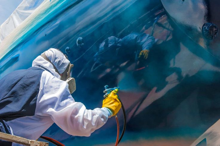 Marine Coating Teknisi mengecat lambung kapal dengan cat epoxy marine coating menggunakan alat semprot, memakai pakaian pelindung lengkap.