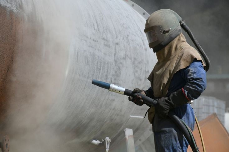 Teknisi sedang melakukan teknik persiapan sandblasting pada permukaan logam besar sebagai bagian dari prosedur awal sandblasting untuk menghilangkan karat dan kotoran